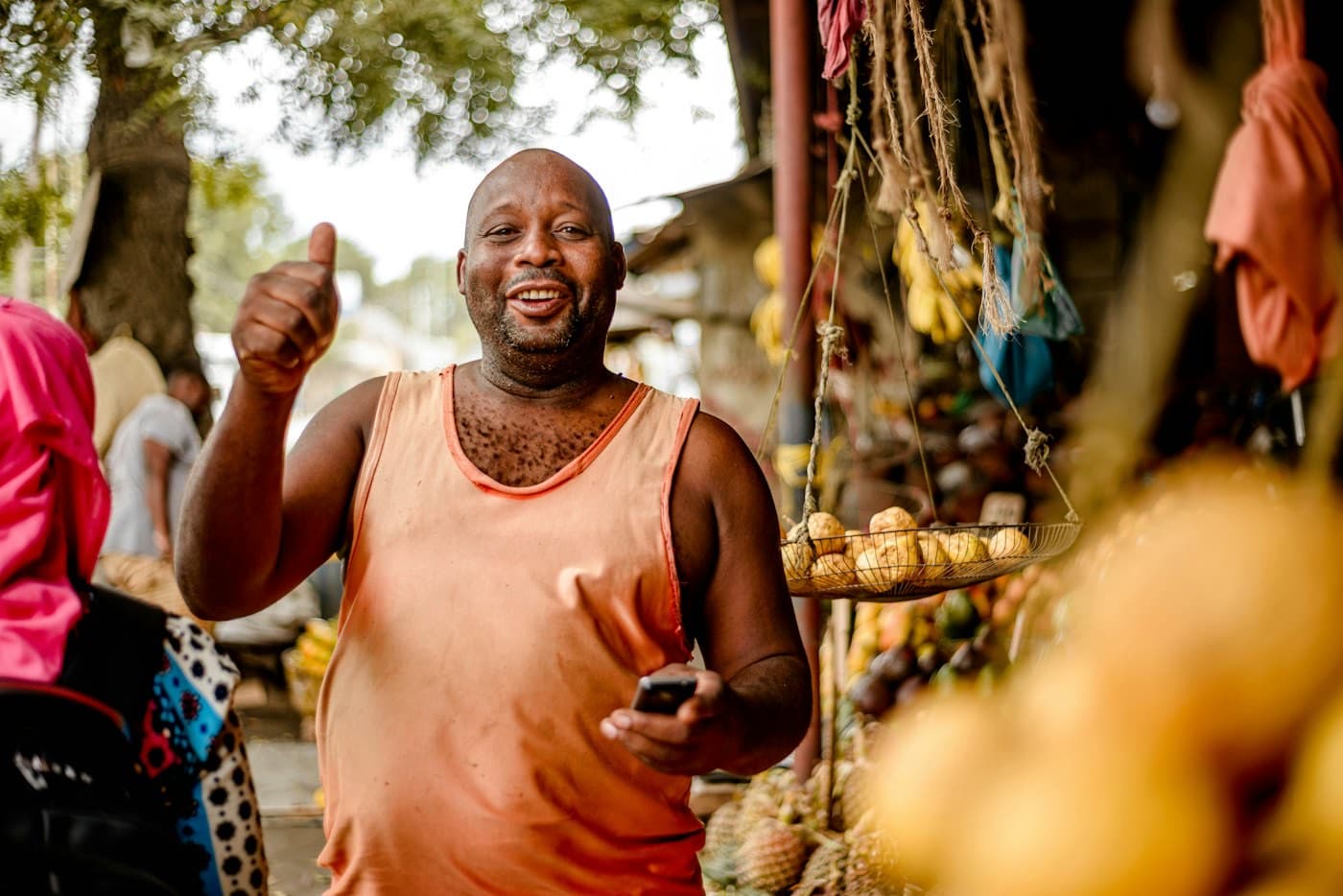A market trader standing at his stall holding a phone, scales hanging behind him